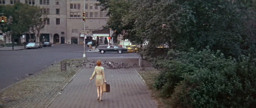 Sweet Charity (1969) movie still: wide — A paved walkway in a city area runs between a dense tree line and road. A woman with a suitcase walks away…