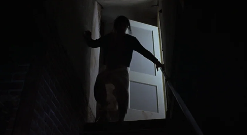 Being John Malkovich (1999) movie still: fullbody — A narrow, dark interior staircase leads down from an open, brightly lit doorway above. A person…