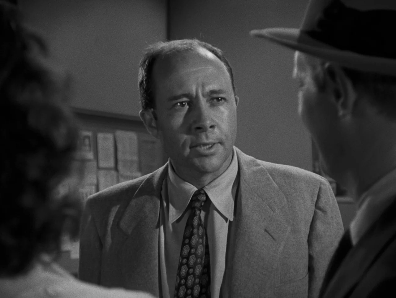 Pickup on South Street (1953) movie still: medium — An interior space features a man speaking to another person in a brimmed hat. A notice board…