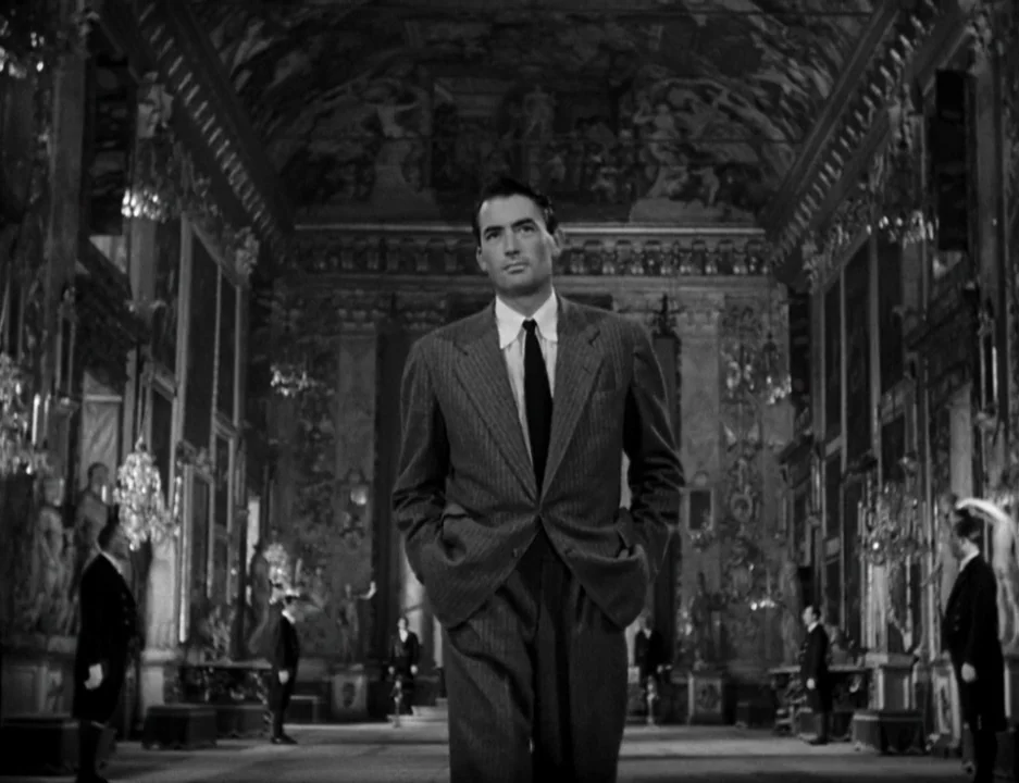 Roman Holiday (1953) movie still: medium — A grand ornate hall features elaborate wall paintings, arched ceilings, and rows of chandeliers. A man…