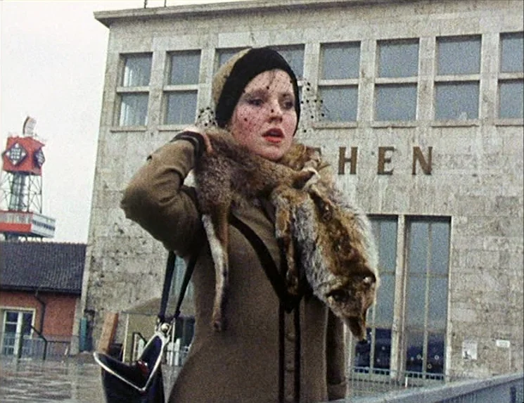 Rio Das Mortes (1971) movie still: medium — A woman stands in front of a modern stone train station building on a day. The woman is wearing a brown…