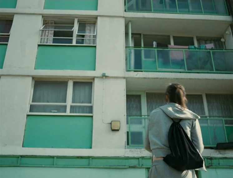 Fish Tank (2009) movie still: fullbody — A multi-story apartment building with white concrete frames and teal panels stands under daylight. A young…