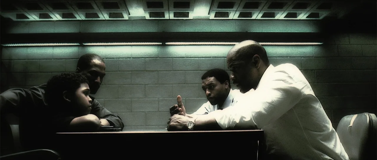 Inside Man (2006) movie still: medium — A stark room features gray concrete block walls and a metal light fixture overhead. Four men sit around a…