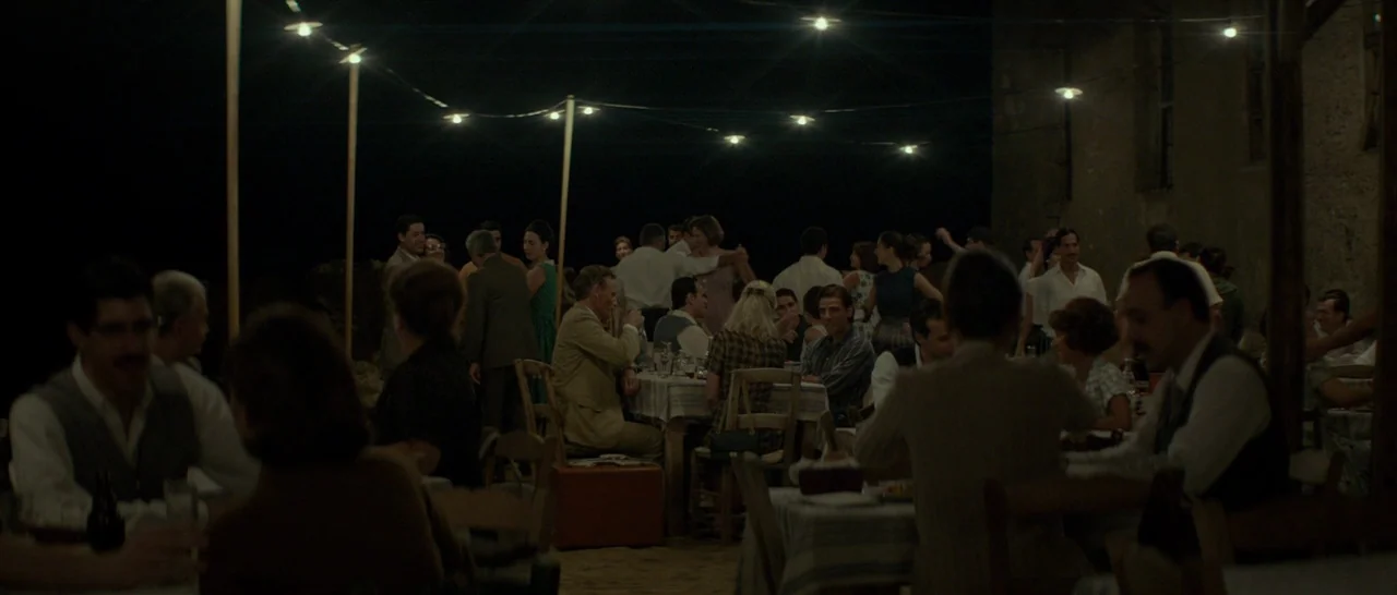 The Two Faces of January (2014) movie still: wide — A night time social gathering of numerous people seated at tables under string lights. Guests…