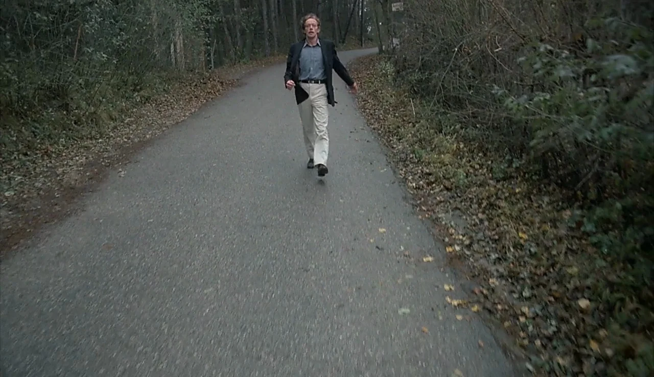 Angst (1983) movie still: fullbody — A man runs toward the camera along an asphalt road bordered by dense trees. Dead leaves cover the road…