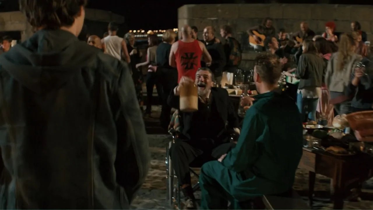 Robot Overlords (2015) movie still: medium — A dark outdoor gathering occurs on a beach with people standing and sitting around tables. An elderly…