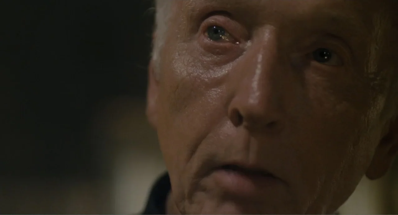 Gates of Darkness (2019) movie still: closeup — A dimly lit indoor space with shadows concealing the background. An elderly man with silver hair…