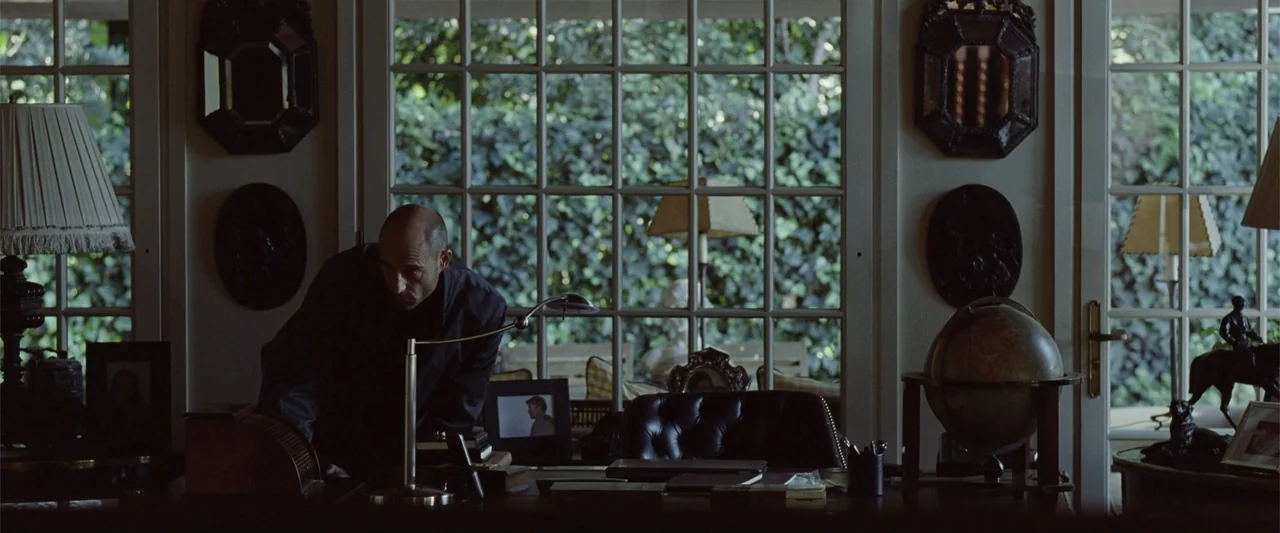 Anna (2013) movie still: medium — A dimly lit room contains a mahogany desk with a globe and framed photographs. A bald man in a dark shirt examines…