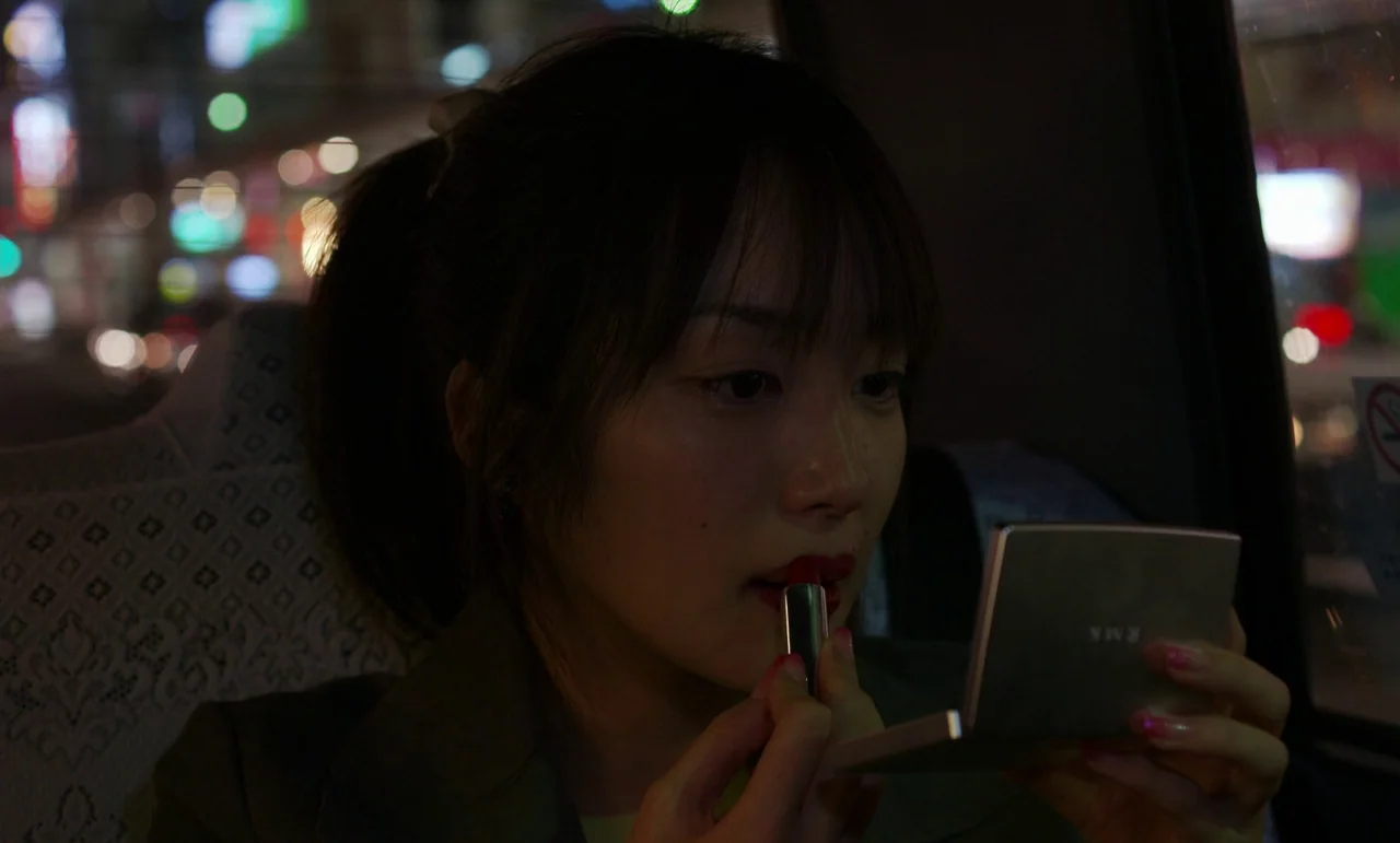 Like Someone In Love (2012) movie still: closeup — The interior of a moving vehicle at night with city lights visible through glass. A young woman…