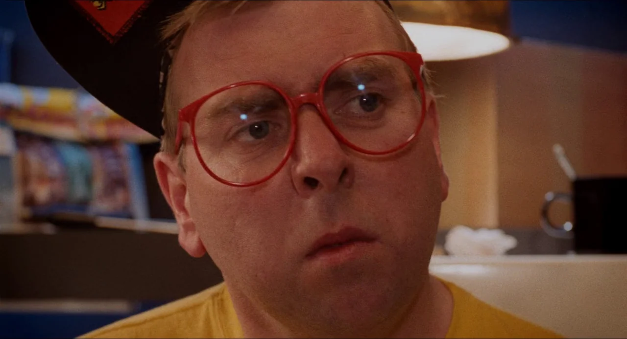 Life Is Sweet (1990) movie still: closeup — A man sits in a booth at a diner with a darkened, out-of-focus background. The man wears bright red…