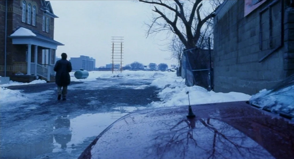 Narc (2002) movie still: wide — A snowy, slush-covered lot sits between a brick building and a cinderblock wall. A man in a dark coat walks away…