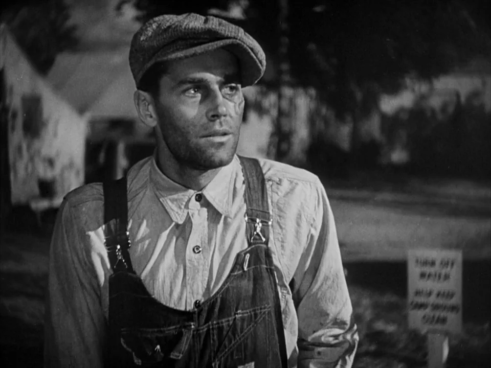 The Grapes of Wrath (1940) movie still: medium — A man in overalls stands in a dimly lit, blurred outdoor nighttime environment. He gazes off-frame…