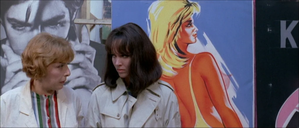 Made in U.S.A. (1966) movie still: medium — A city street featuring large, colorful advertising posters attached to a wall. Two women stand in front…