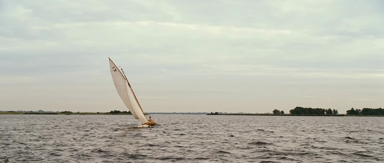 Black Book (2006) movie still: wide — An open lake under a flat, grey overcast sky with distant tree-lined shores. A single small wooden sailboat…