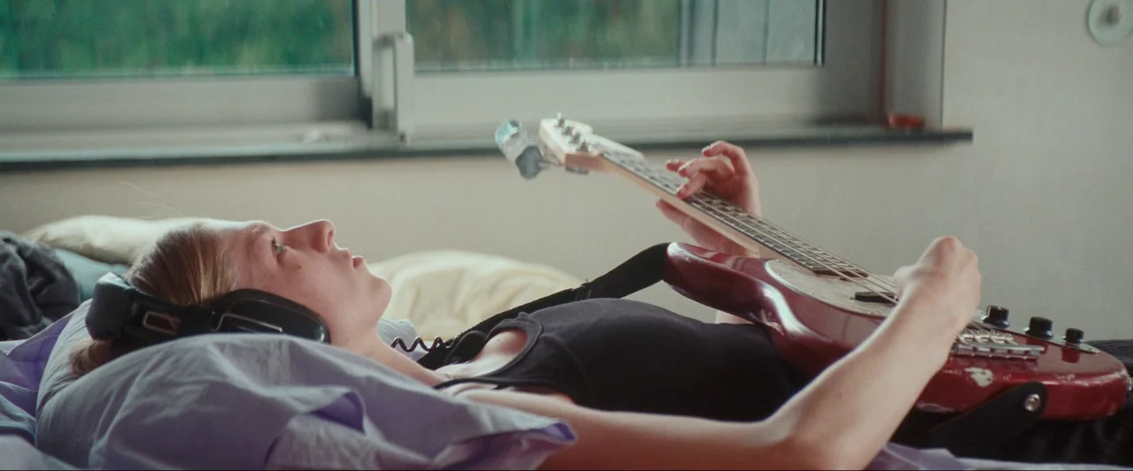 Cuckoo (2024) movie still: medium — A person lies on a bed in a room with a large window. The subject is wearing black headphones and playing a red…