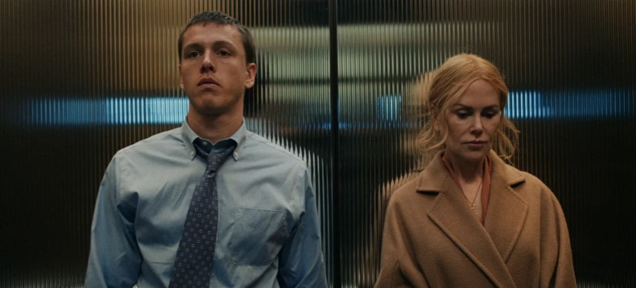 Babygirl (2024) movie still: medium — An elevator interior features dark, vertically ribbed glass walls behind two individuals. A man in a blue…