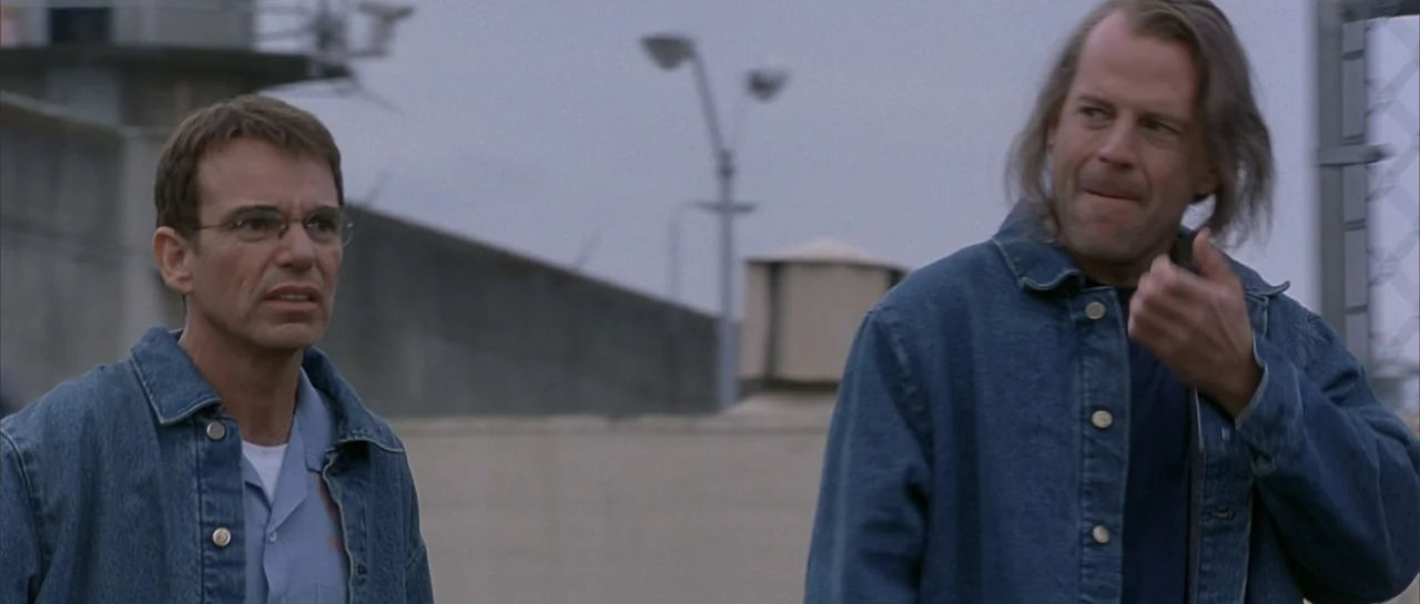 Bandits (2001) movie still: medium — A secure outdoor area with a high gray concrete wall and a metal fence. Two men stand together in the…