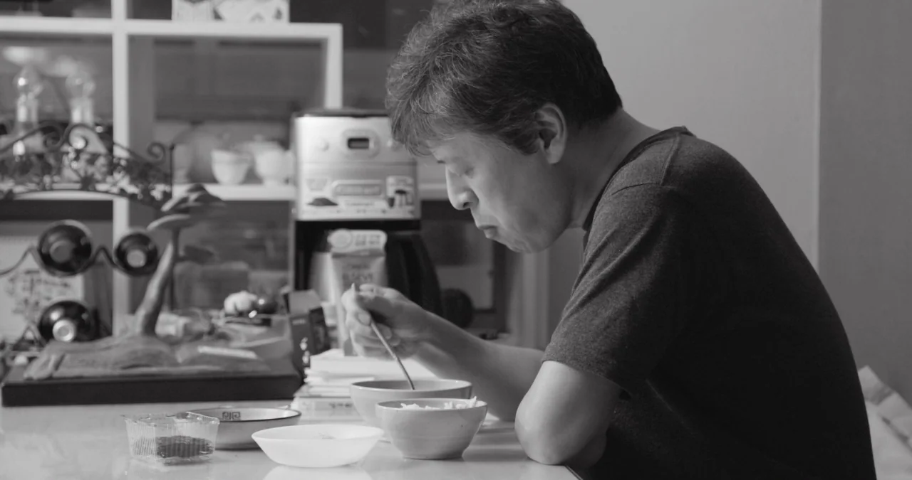 The Day After movie still: medium — A man sits at a dining table with bowls and a coffee machine visible. He eats with a spoon while leaning on the…