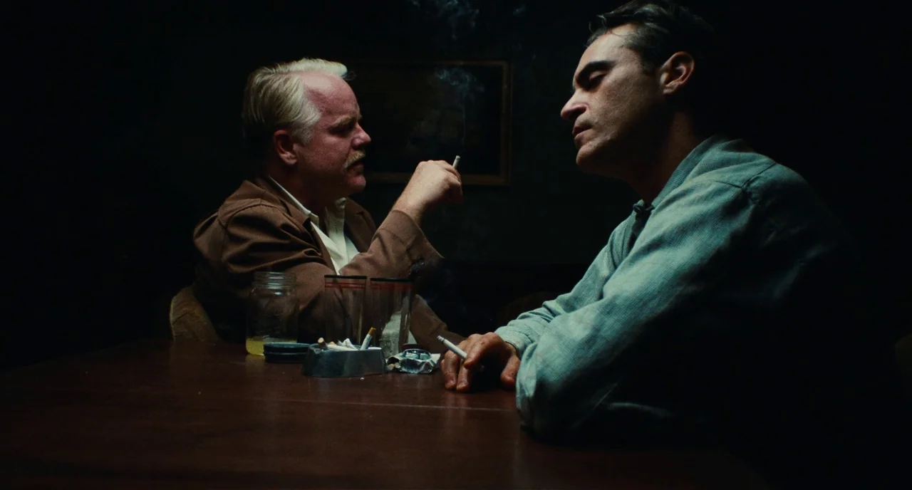 The Master (2012) movie still: medium — A dimly lit, sparse interior setting with a dark wooden table as the focus. Two men sit opposite each other…