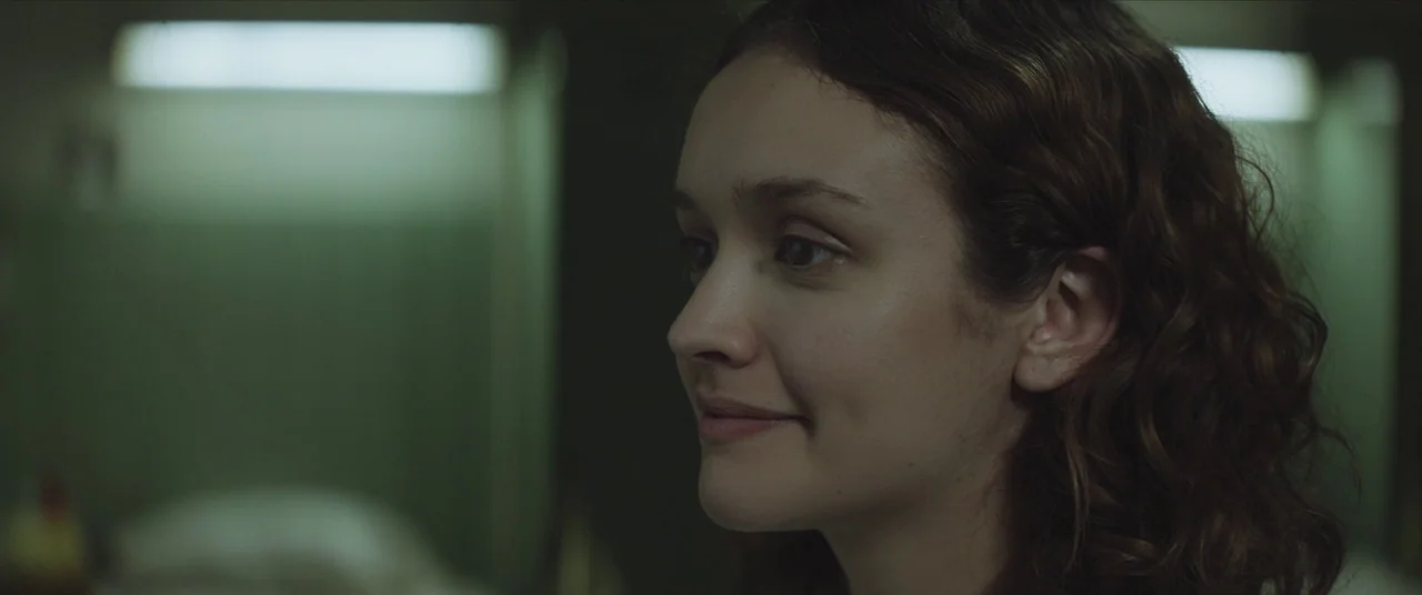 Thoroughbreds (2017) movie still: closeup — A dimly lit room with blurred overhead light fixtures against muted green walls. A young woman with wavy…