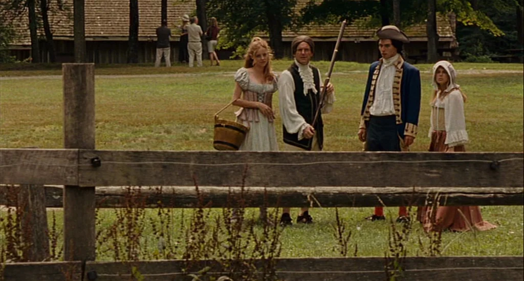 Choke (2008) movie still: wide — Four people stand in a grassy field behind a split-rail wooden fence. A long wooden barn is visible in the…