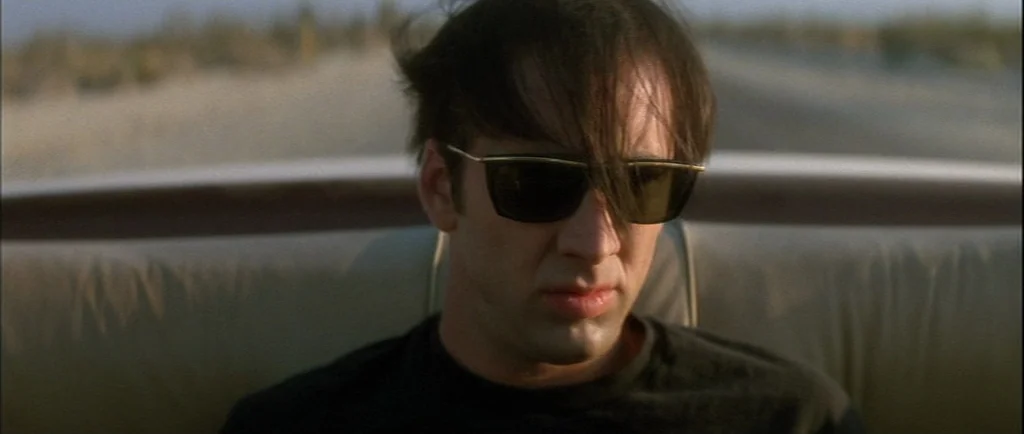 Wild At Heart (1990) movie still: closeup — A man sits in the passenger seat of an open convertible moving through desert. The background is a blur…