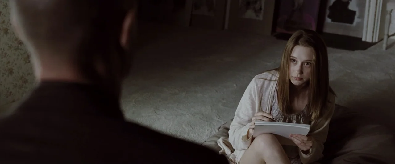 Anna (2013) movie still: medium — A dimly lit room with carpeted floors and wallpaper. A woman with long brown hair sits on the floor writing in a…