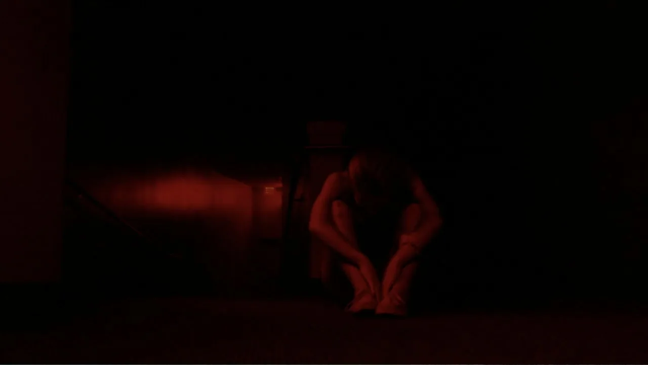 The Gallows (2015) movie still: medium — A dark staircase landing is illuminated by a single red light source. A person sits curled on the ground…