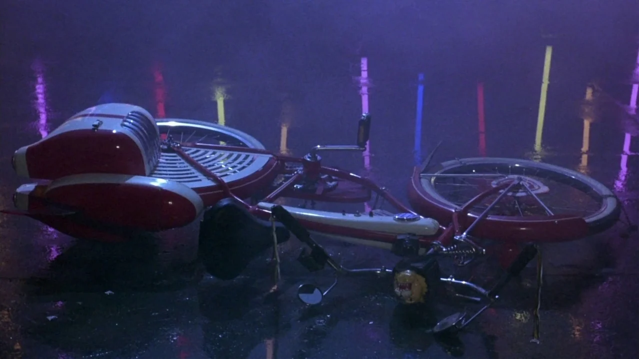 Pee-Wee’s Big Adventure (1985) movie still: medium — A red and white stylized vintage bicycle lies on wet, reflective dark pavement at night. The…