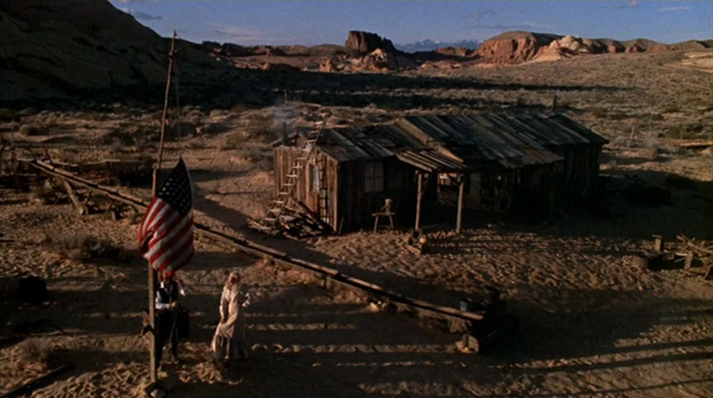 The Ballad of Cable Hogue (1970) movie still: wide — A remote wooden cabin sits in a sprawling, arid desert landscape with rocky outcrops. A man and…
