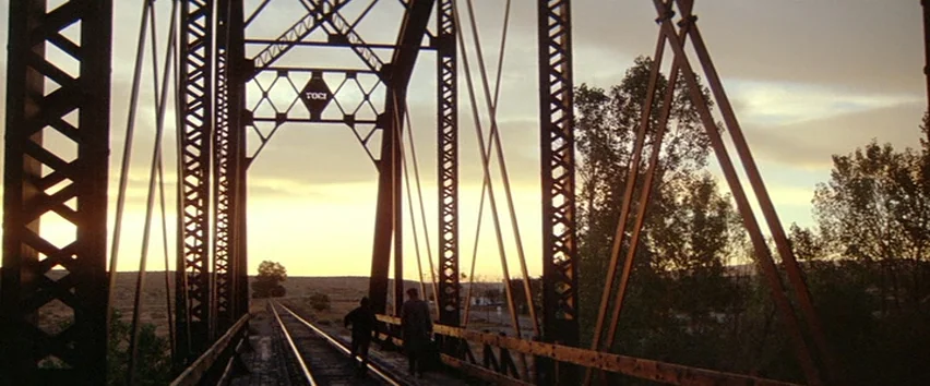 Scarecrow (1973) movie still: wide — A metal truss railway bridge spans over a landscape at sunset. Two figures walk away from the camera along the…