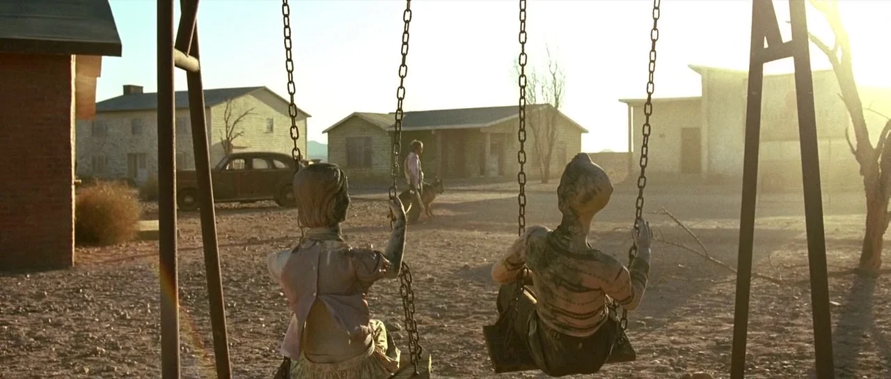 The Hills Have Eyes (2006) movie still: wide — An arid, dusty ghost town features old wooden buildings under a bright, hazy sky. Two weathered…
