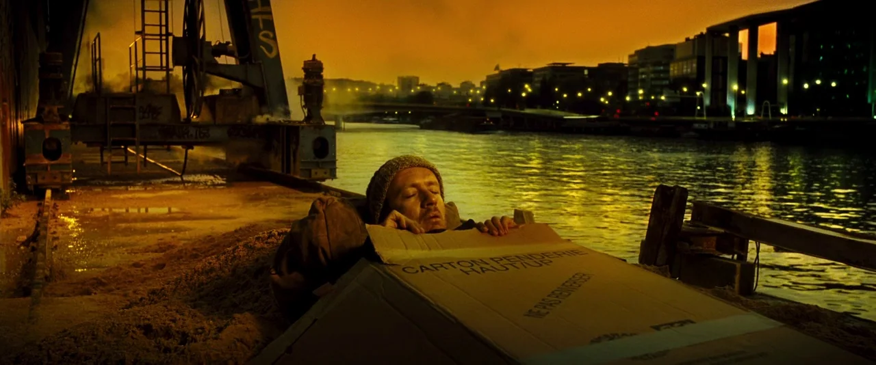 Micmacs (2009) movie still: medium — A man lies inside a large cardboard box on a sandy riverbank at dusk. Massive industrial crane structures rise…