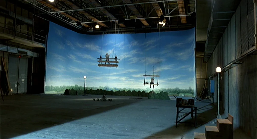 Intervista (1987) movie still: wide — A massive soundstage features a large curved backdrop painted with a blue sky landscape. Two workers are…