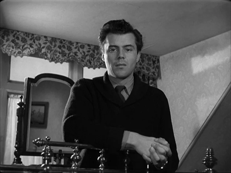 The Sleeping Tiger (1954) movie still: medium — A man stands in a bedroom interior with patterned wallpaper and a vanity mirror. He leans his…