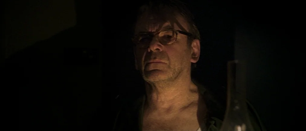 Time Of The Wolf (2003) movie still: closeup — An older man stands in a dimly lit, undefined dark space. He faces forward while illuminated by a…