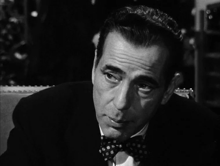 In a Lonely Place (1950) movie still: closeup — A dimly lit indoor space with the subject positioned in the mid-foreground. A man in a suit and bow…