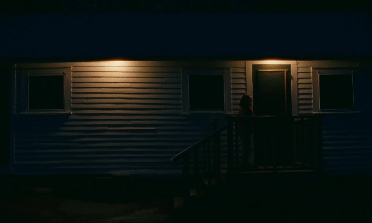 Janet Planet (2023) movie still: wide — A white-sided house wall at night features three dark windows and a lit doorway. A lone person stands on the…