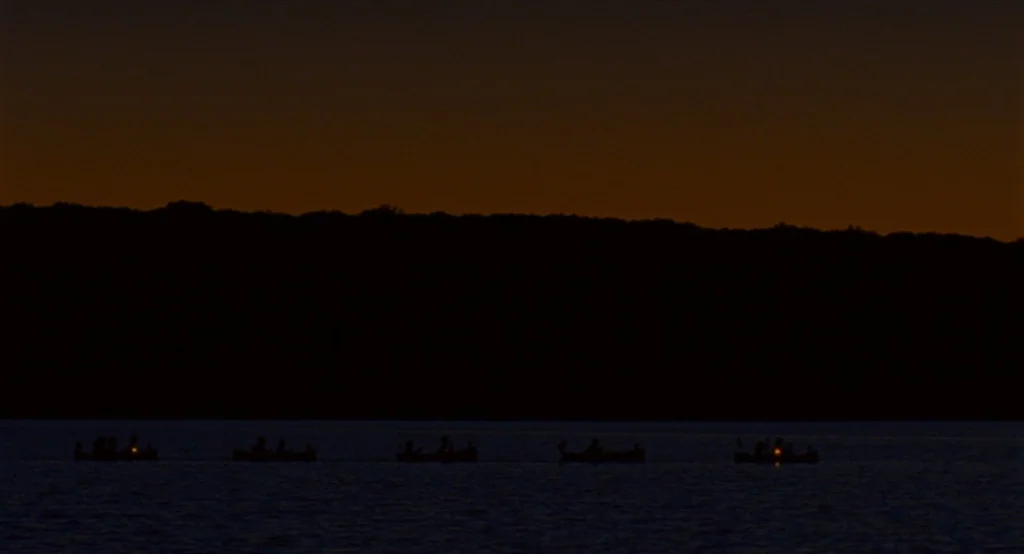 Moonrise Kingdom (2012) movie still: wide — A dark, calm lake surface stretches before a solid, black silhouette of a forested shoreline. Five small…