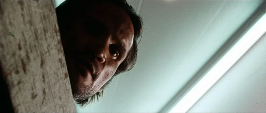 Messiah of Evil (1973) movie still: closeup — An interior space viewed from a low angle under harsh, clinical lighting conditions. A man's face…