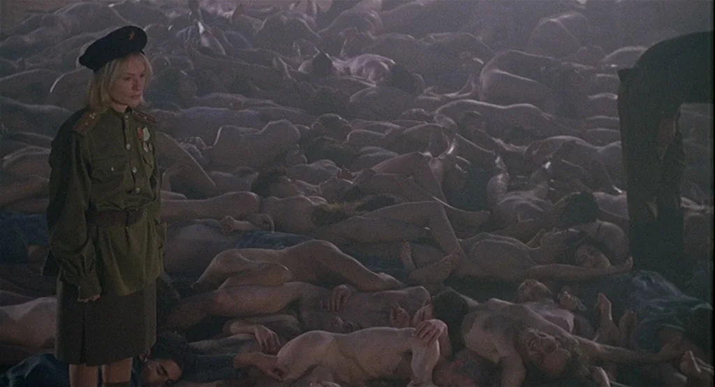 Deux (2002) movie still: fullbody — A dimly lit warehouse floor is covered with a large pile of nude bodies. A woman in a Soviet military uniform…