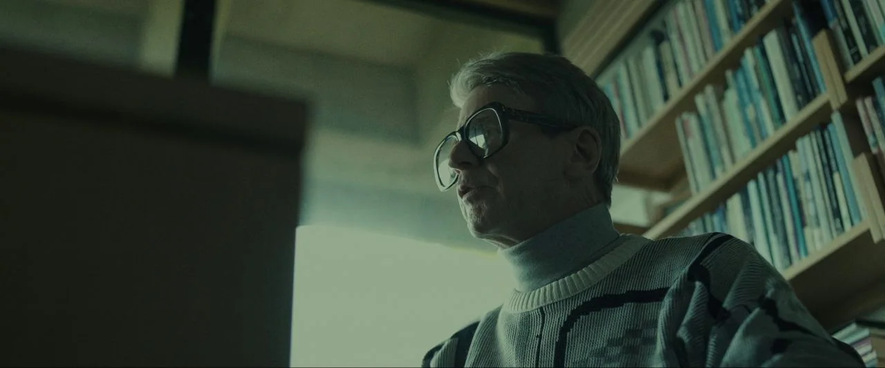 Come True (2020) movie still: medium — A man stands in a room with shelves filled with books. He looks toward a computer screen positioned off-camera.