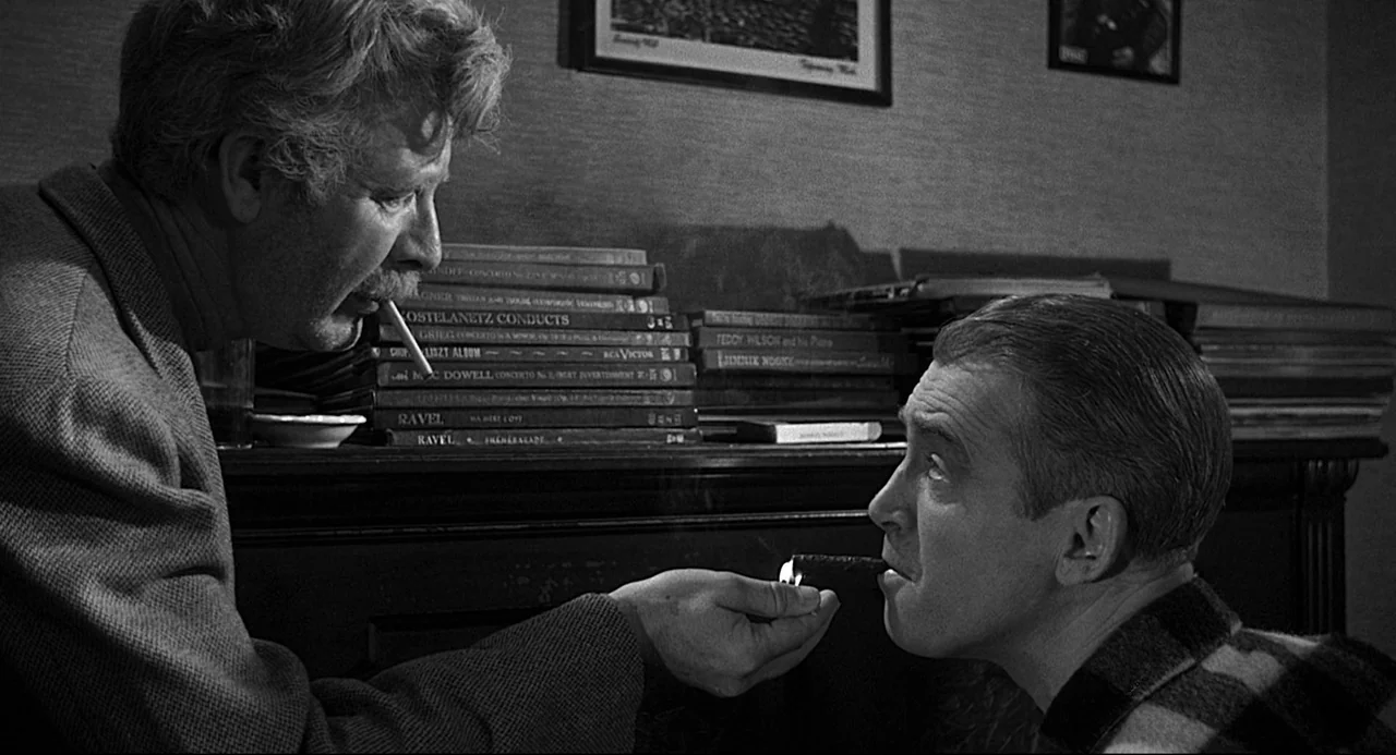 Anatomy of a Murder (1959) movie still: medium — A dark, wood-paneled room is filled with stacks of phonograph records on a sideboard. An older man…