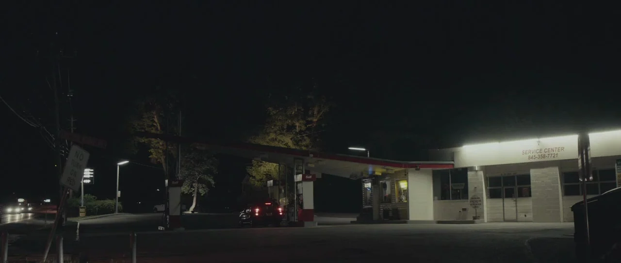 Meadowland (2015) movie still: wide — A nighttime view of a quiet gas station and service center with bright lighting. A lone vehicle sits under the…