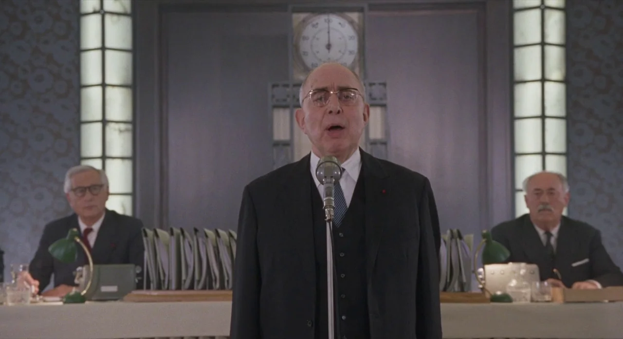 Populaire (2012) movie still: medium — A formal hearing room features a central desk with a large analog clock behind. A man in a dark suit stands…