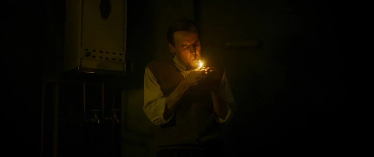 Earwig (2022) movie still: medium — A dark, spartan interior contains a metallic wall-mounted water heater in the background. A thin man in a…