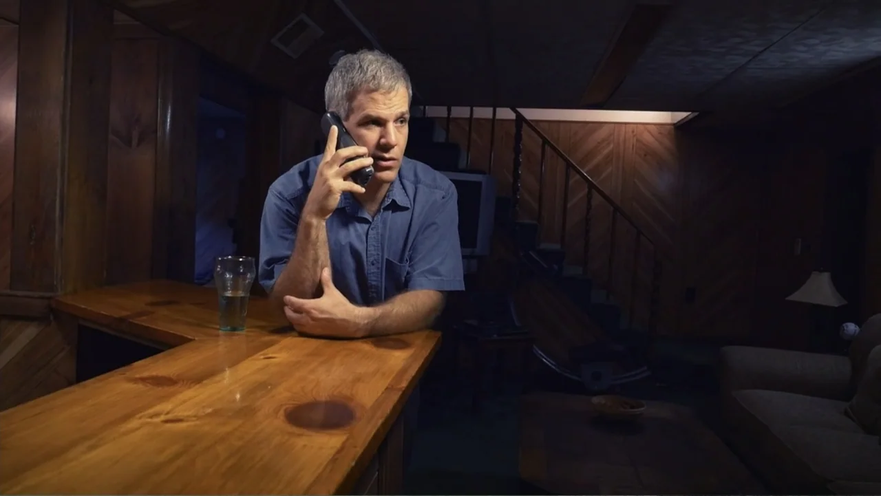 Beware of Angels (2016) movie still: medium — A dark finished basement features wood-paneled walls and a built-in wooden bar counter. A middle-aged…