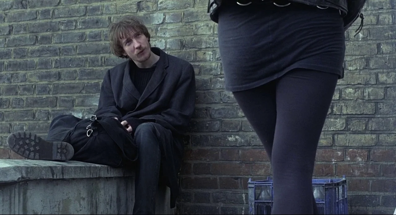 Naked (1993) movie still: medium — A man sits against a weathered yellow brick wall outdoors near a plastic crate. A woman wearing black stands in…