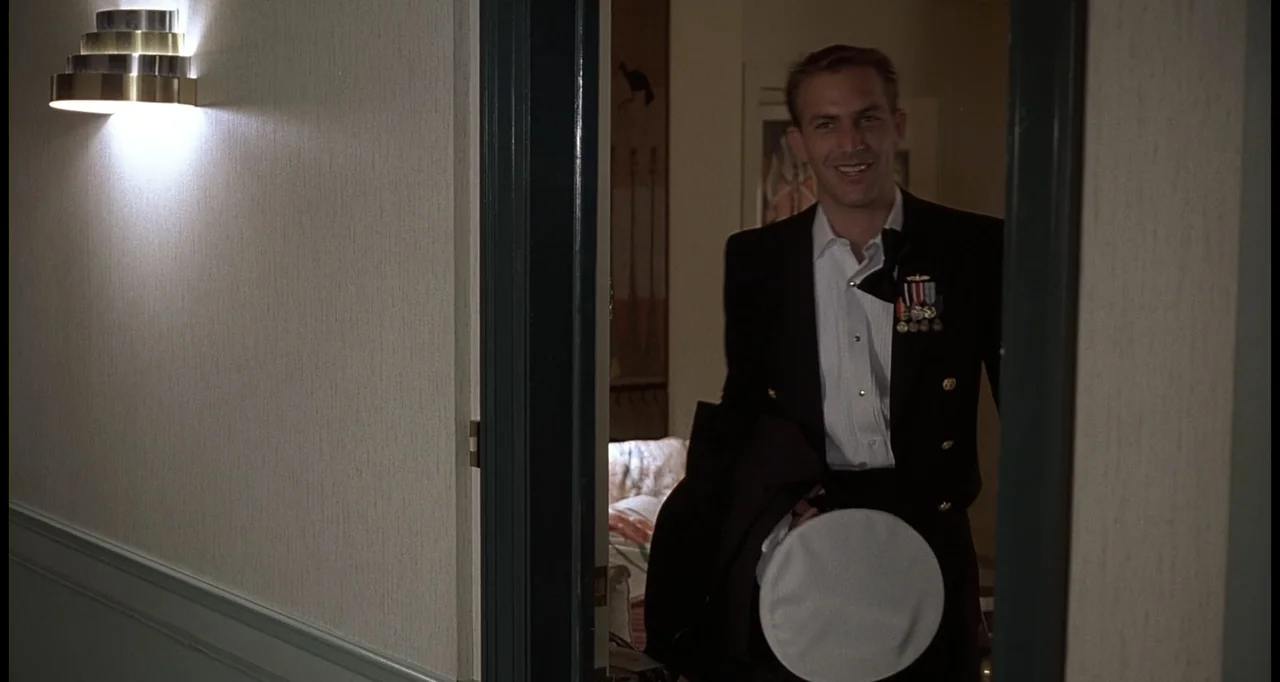 No Way Out (1987) movie still: medium — A well-lit hallway with light walls features a dark doorframe leading into a room. A man in a naval dress…