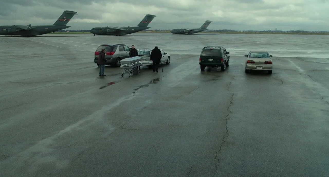 Last Flag Flying (2017) movie still: wide — A wet concrete airfield runway features three large cargo airplanes parked in the distance. Three men…